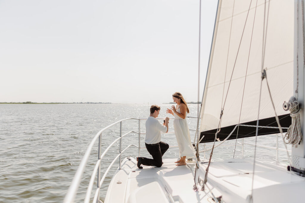 A pair standing together on a sailboat, celebrating their romantic surprise proposal.
