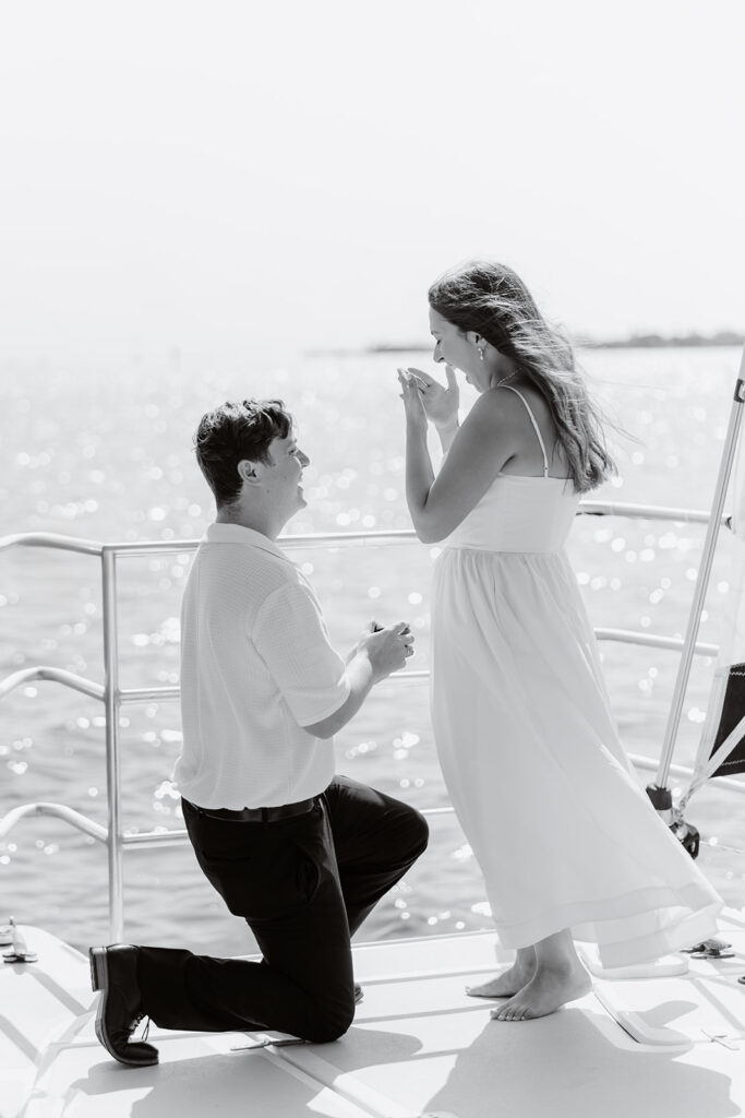 A pair standing together on a sailboat, celebrating their romantic surprise proposal.
