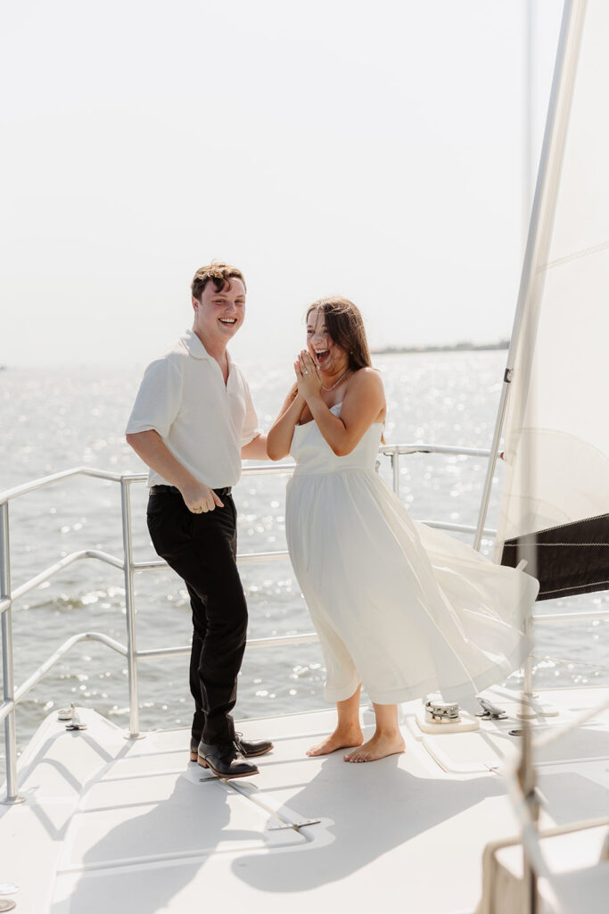 A pair standing together on a sailboat, celebrating their romantic surprise proposal.
